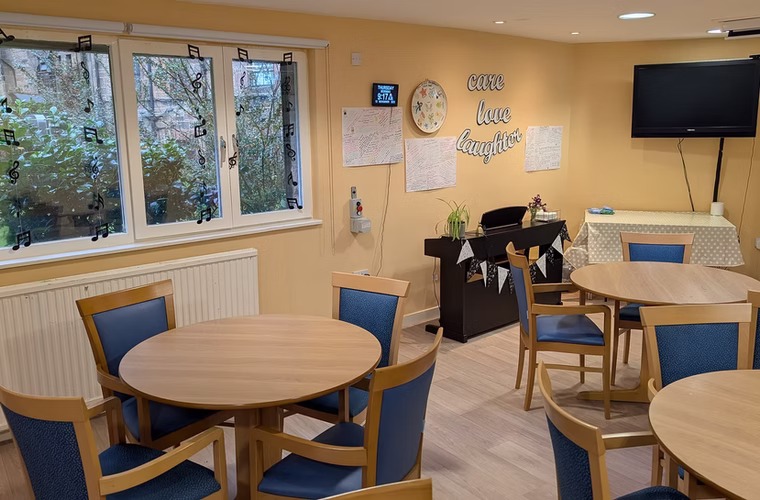 The bright club room at The Open Door with round tables, comfortable chairs and a piano.