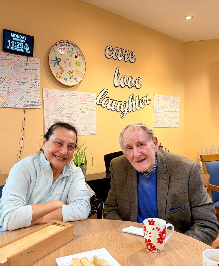 A volunteer and a member sharing a smile over a cup of tea at The Open Door, beneath the club's 'care, love, laughter' wall.